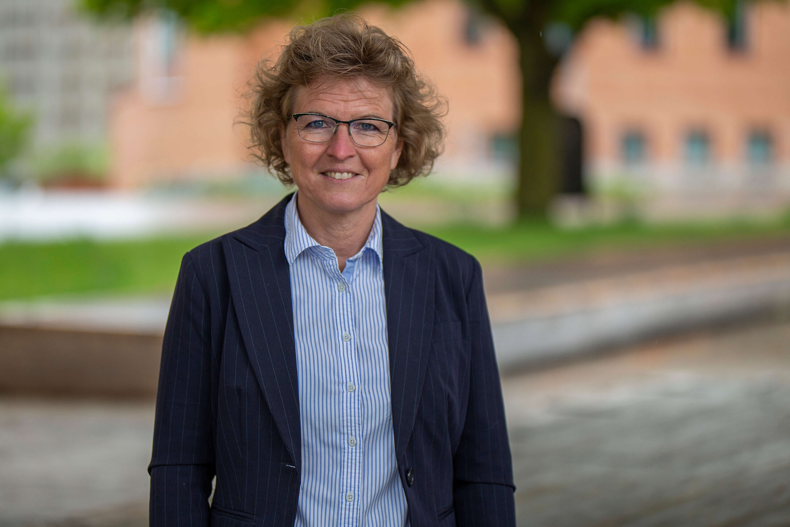 Porträtt på kvinna med kort ljust hår, glasögon, ljusblå skjorta och mörkblå kavaj som står utomhus