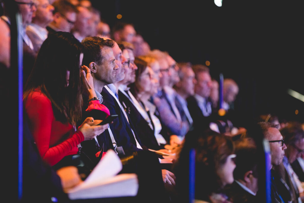 En grupp människor sitter tätt tillsammans i en publik på ett evenemang inomhus. De flesta är fokuserade framåt, medan en rödklädd kvinna i förgrunden tittar på sin telefon. Belysningen är mjuk och stämningen är uppmärksam.